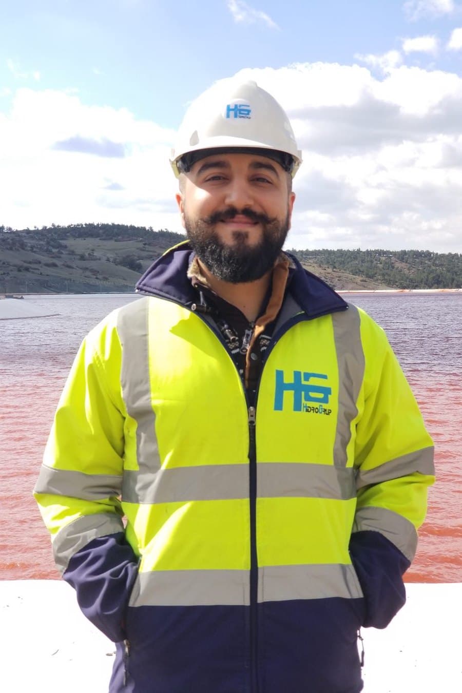 In a hard hat and hi-vis jacket on site, a pink pond in the background